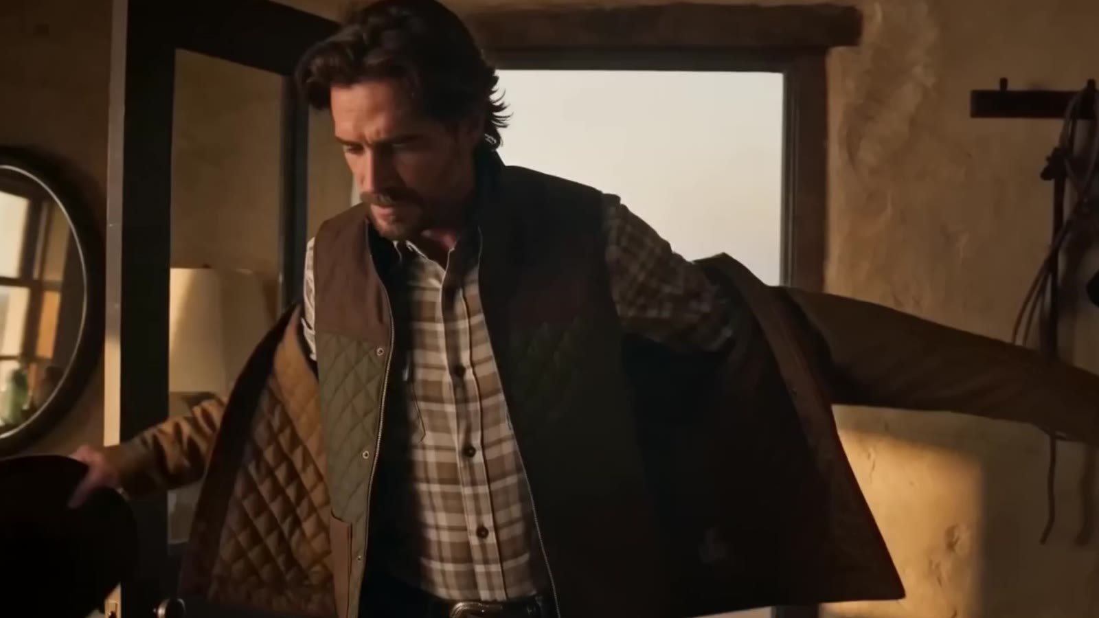 Man pulling on a quilted vest in a tack room